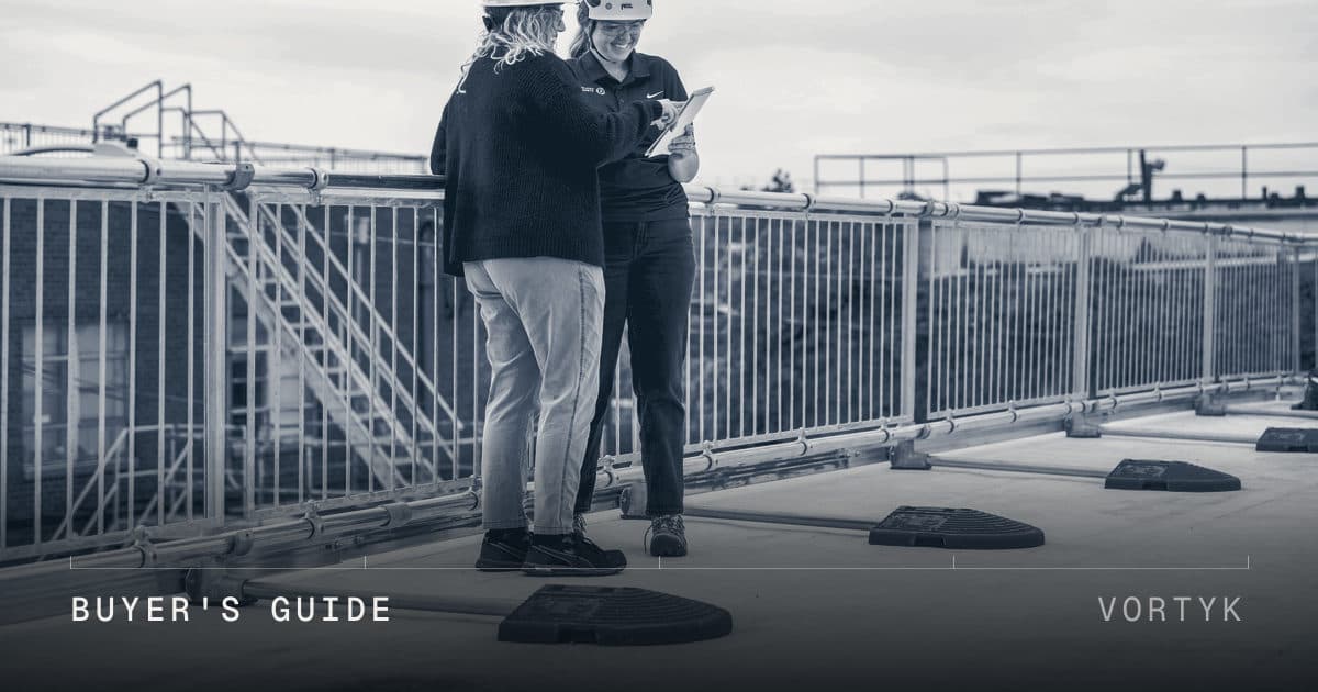 Two inspectors in hard hats reviewing a tablet together on a flat roof, with guardrails and rooftop equipment behind them.